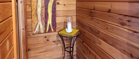 Interior de una cabaña de madera, con un muro de madera, un cuadro enmarcado, una mesita con una vela y suelo de madera.