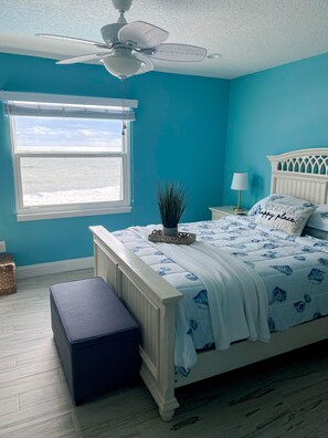 A bedroom with a bed, a ceiling fan, a window with a view, and a nightstand with a lamp.