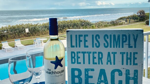 A bottle of wine, two wine glasses, and a sign with the text "Life is simply better at the beach" on a table with a view of the ocean.