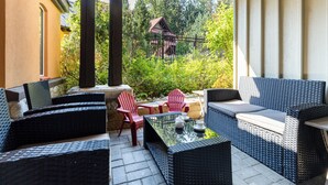 An outdoor patio with wicker furniture, a glass-top table, and a view of a house surrounded by trees.