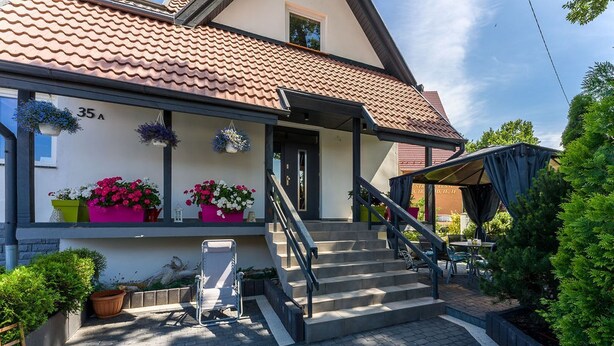 A two-story house with a tiled roof, a front porch with steps, and potted plants on the balcony.