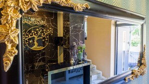 A room with a large mirror reflecting a marble wall, a gold-framed mirror, and a small vase with purple flowers.