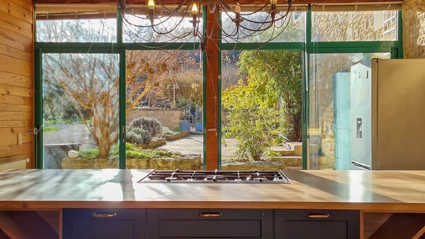Une cuisine dotée d’un îlot central, d’un plafond en bois et de grandes fenêtres donnant sur un jardin.