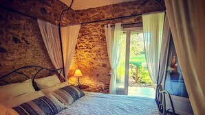Une chambre avec un mur en pierre, un lit avec une literie à motifs, une lampe de chevet et une vue sur un jardin, visible à travers une porte vitrée.
