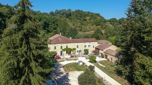 Un bâtiment traditionnel en pierre, avec un toit recouvert de tuiles, entouré d’une végétation luxuriante et surplombé d’un ciel bleu azur.