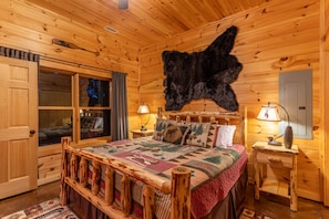 A cozy log cabin bedroom with a large fur rug, a wooden bed with a quilt, two bedside tables with lamps, and a sliding glass door.
