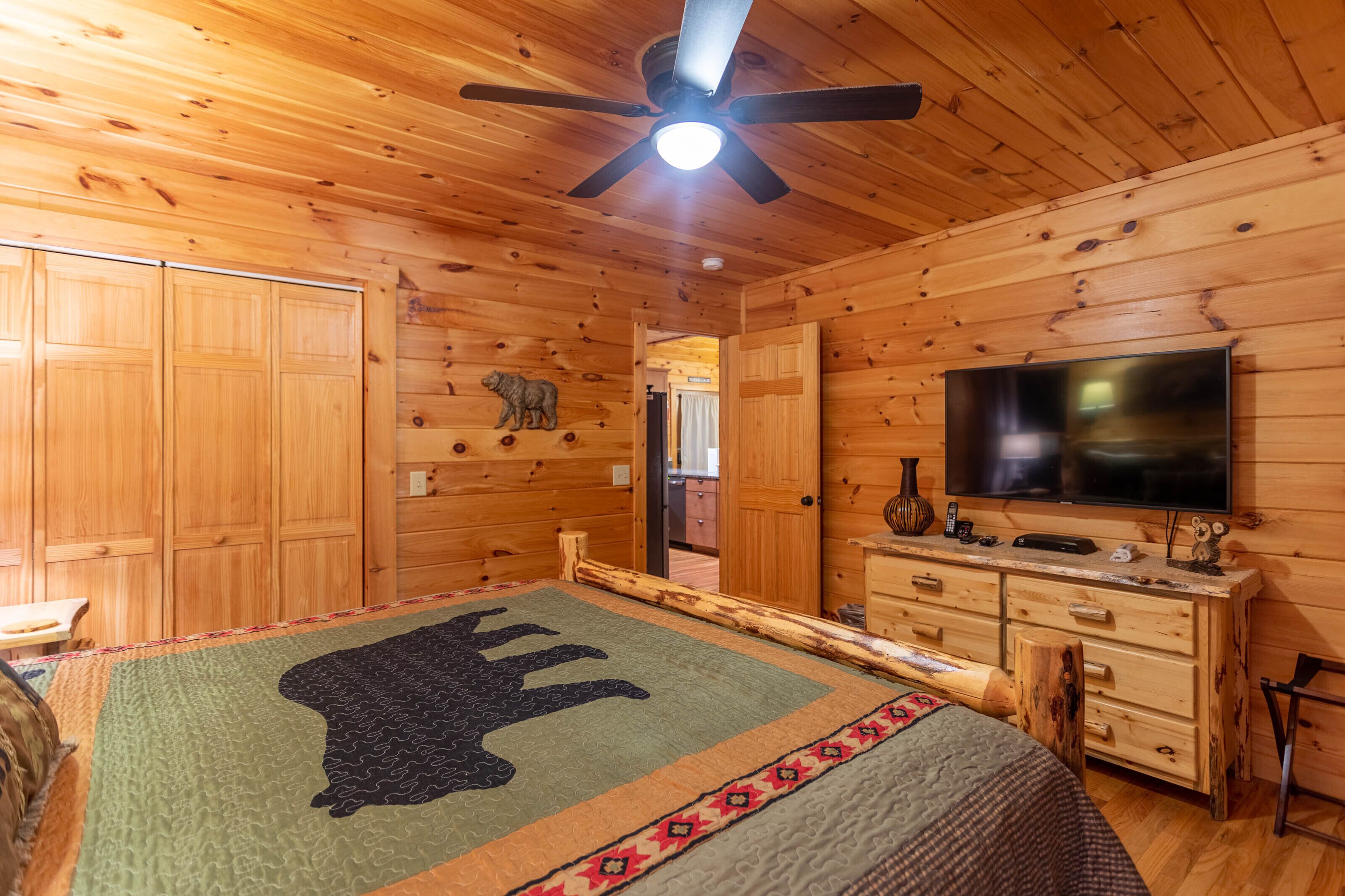 A log cabin bedroom with a large bed, wooden walls, a ceiling fan, a television, and a dresser.