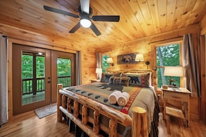 A wooden cabin bedroom with a bed, two bedside lamps, a ceiling fan, and a window with a view of trees.