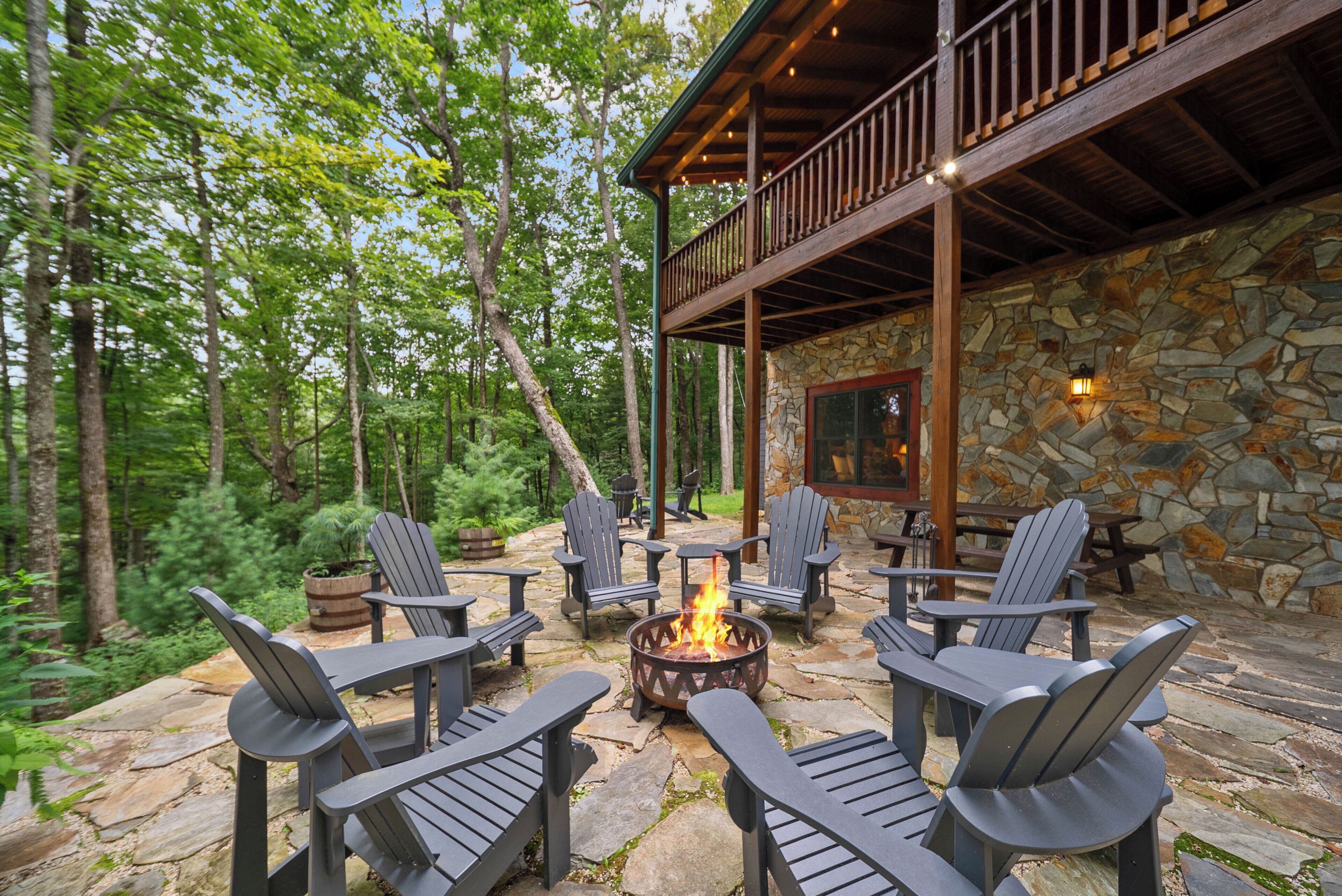 Un espace extérieur aménagé avec des chaises Adirondack disposées autour d’un foyer, le tout adossé à un mur de pierres et entouré d’arbres.