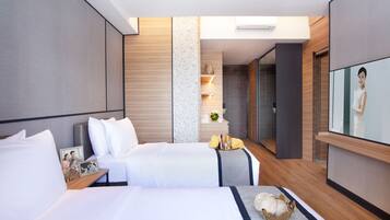 Habitación de hotel con dos camas, televisión, una mesita con un cuenco y un marco de fotos, y un baño visible a través de una puerta de vidrio.