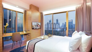 Une chambre d’hôtel avec un grand lit, un bureau et une chaise. De grandes fenêtres offrent une vue sur le paysage urbain.