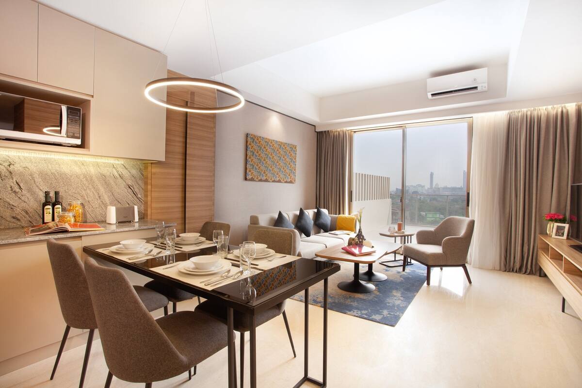 Una habitación de hotel moderna con comedor, un conjunto de sofás, televisor y un amplio ventanal con vista a la ciudad.