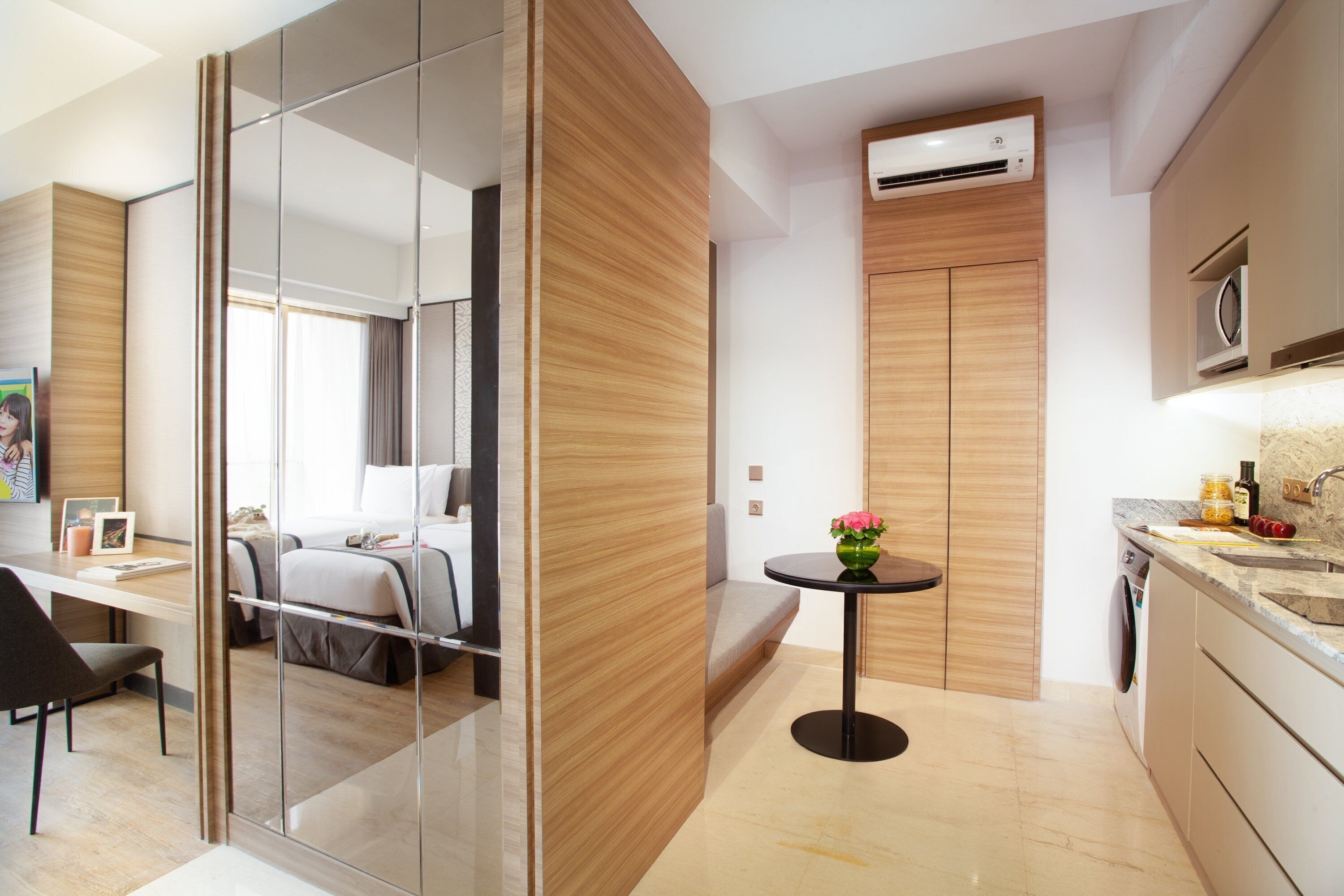 A modern hotel room with a bed, a desk, a chair, a small table with a vase, and a kitchen area with a microwave and sink.