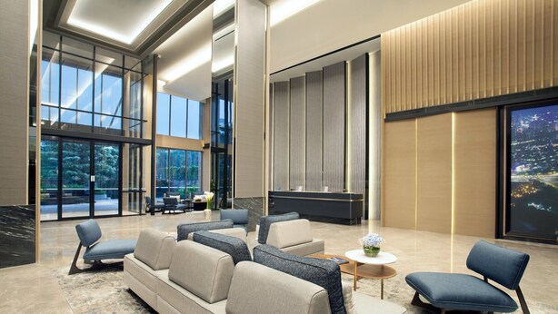 Un hall d’entrée moderne doté d’un canapé, de fauteuils, d’un comptoir de réception et de grandes fenêtres offrant une vue sur l’extérieur.