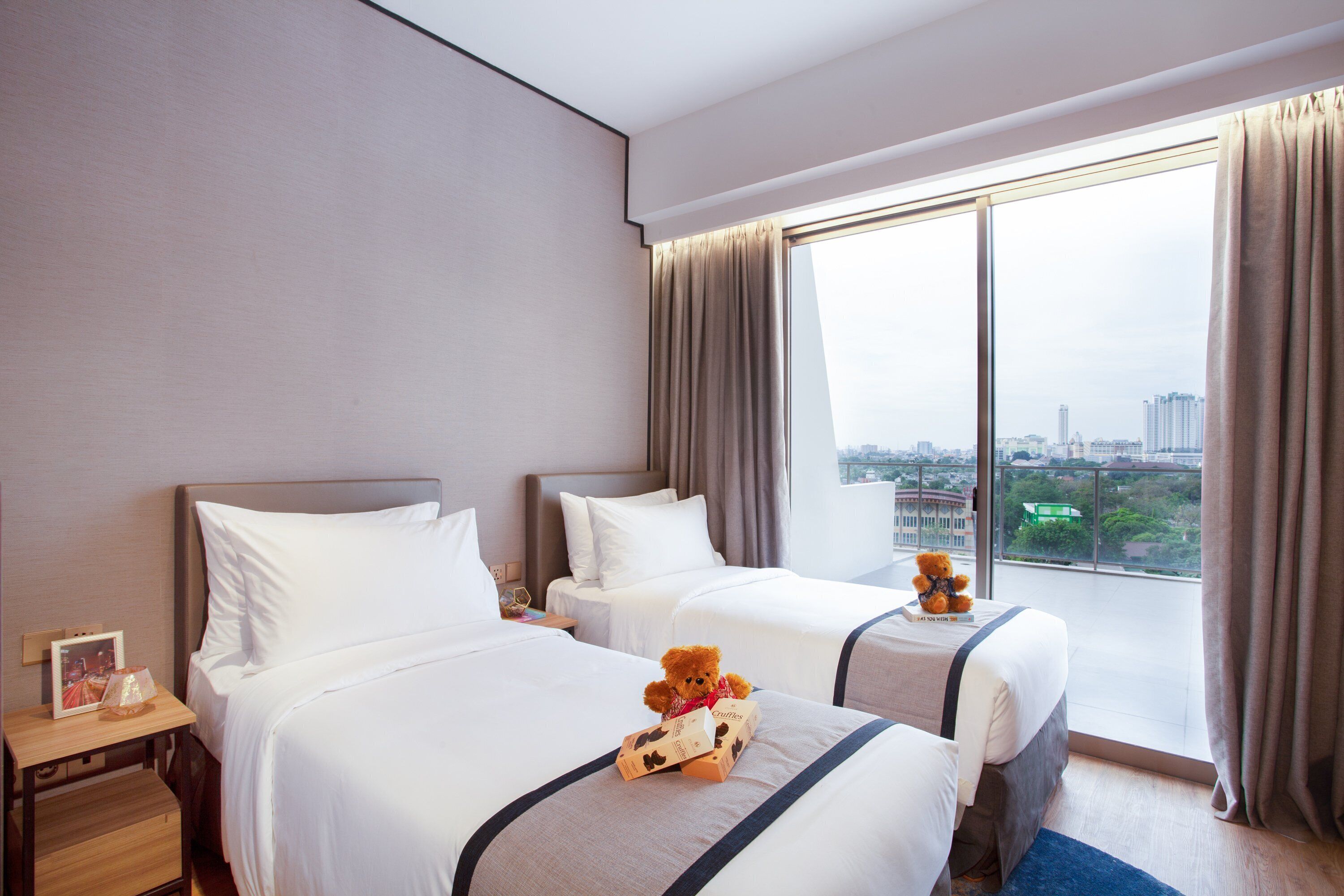 Ein Hotelzimmer mit zwei Betten, einem großen Fenster mit Vorhängen und einem Teddybären auf einem Bett.