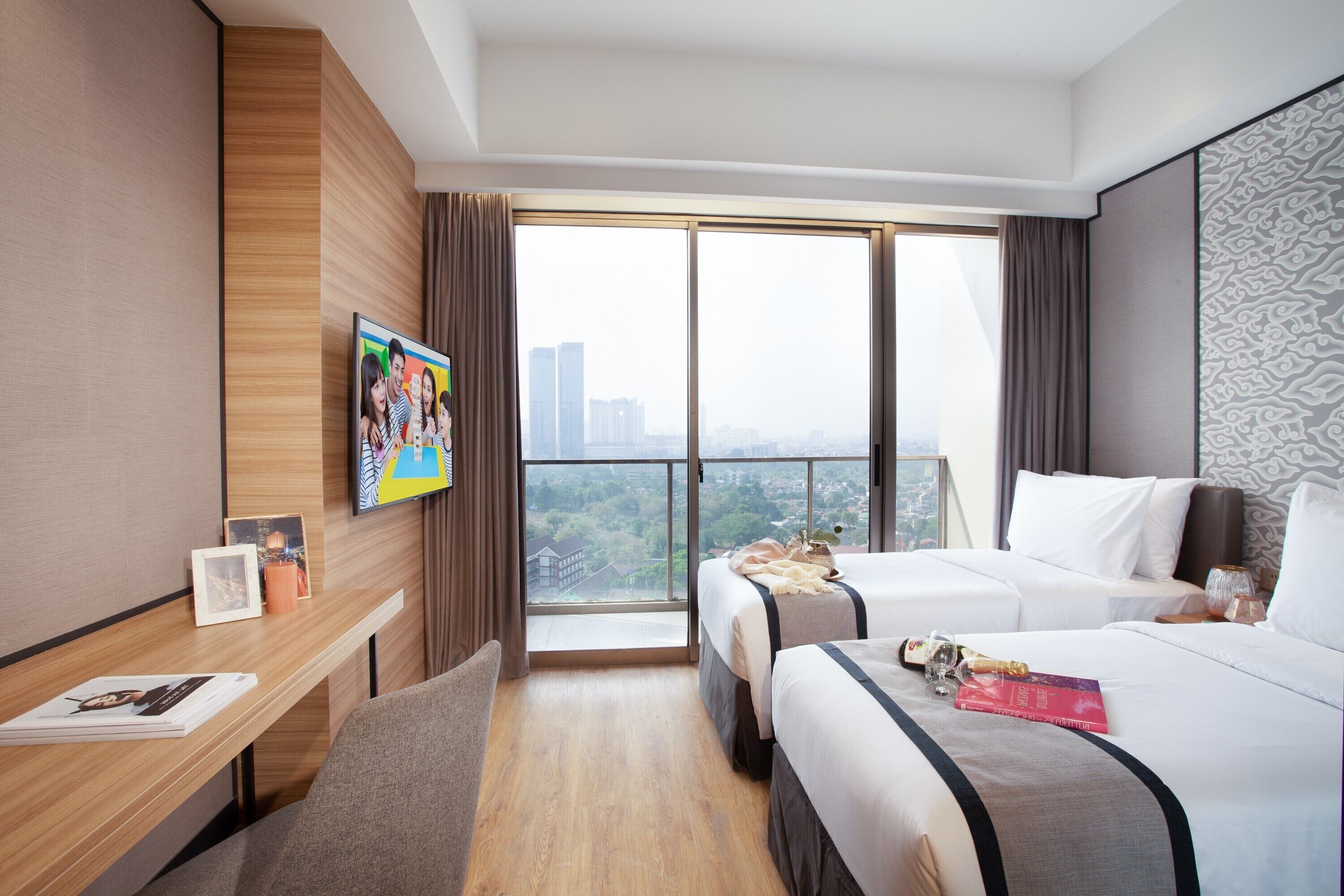 Habitación de hotel con dos camas, un escritorio, televisor y un ventanal grande con vista a la ciudad.