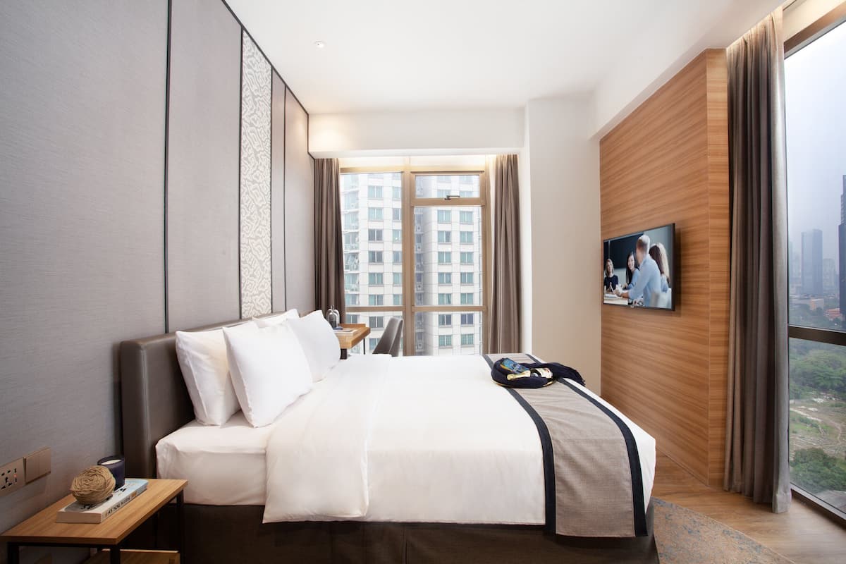 Habitación de hotel con una cama grande, un televisor de pantalla plana montado en la pared, un muro decorativo de madera y vistas a un paisaje urbano a través de la ventana.