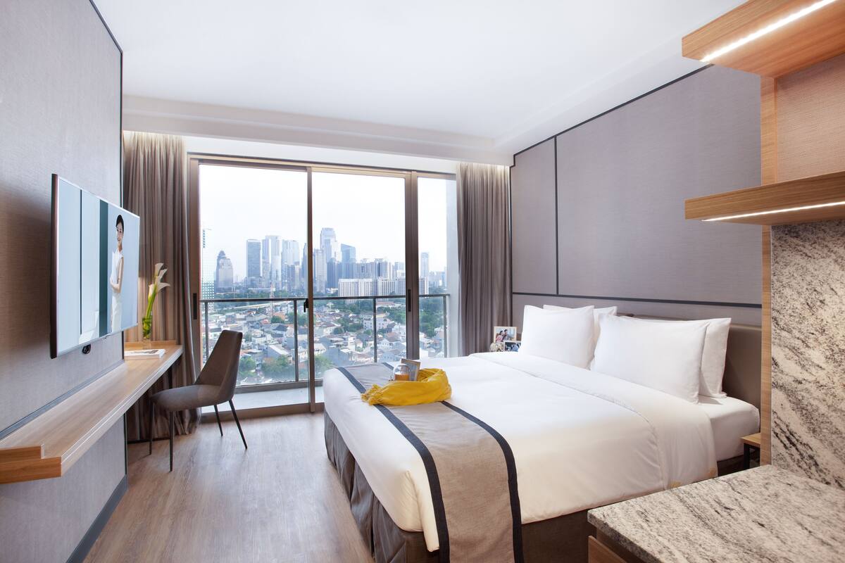Una habitación de hotel moderna con una cama grande, un escritorio y vistas de la ciudad a través de amplios ventanales.