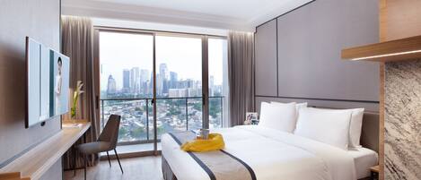 A modern hotel room with a large bed, a desk, and a view of the cityscape through large windows.