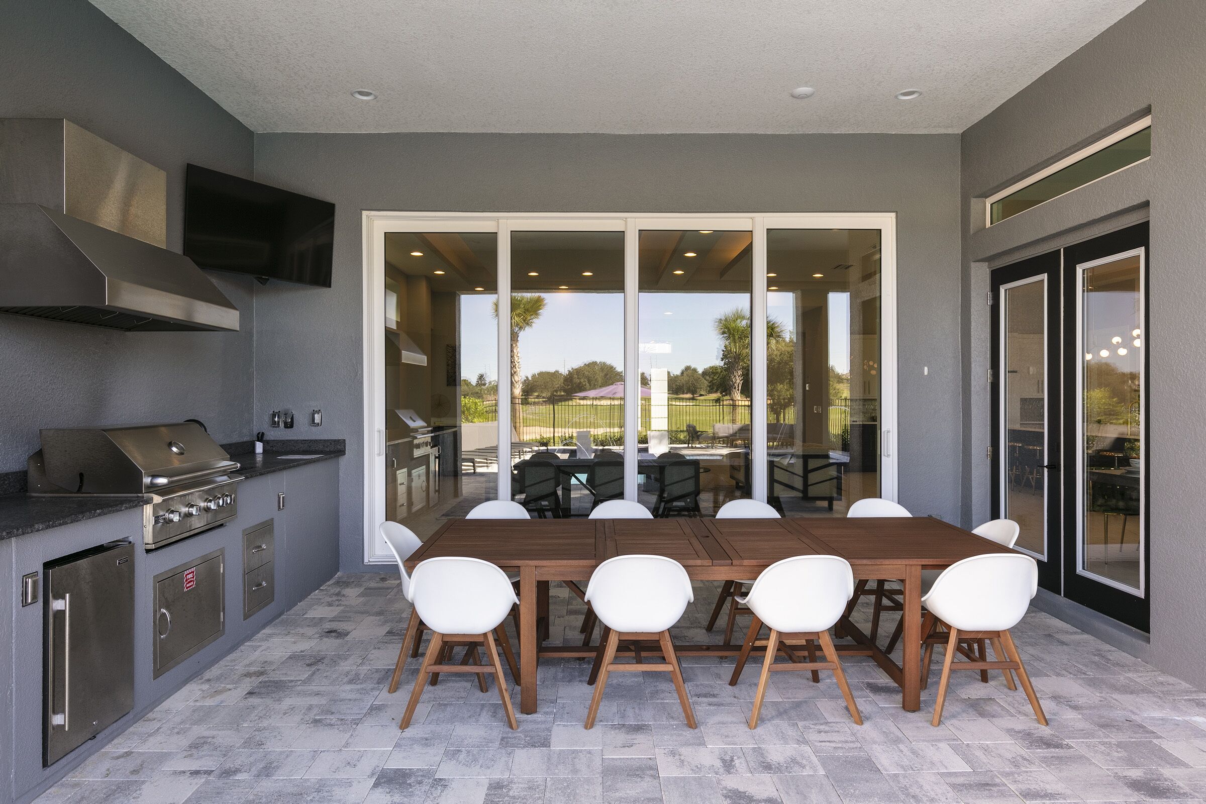 Une cuisine extérieure équipée d’un barbecue, d’une table à manger et de chaises, donnant sur un patio couvert avec vue sur l’extérieur.
