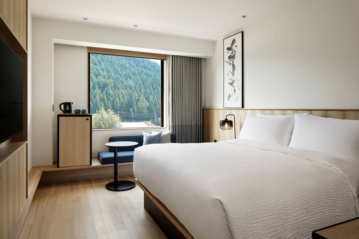 A modern hotel room with a large bed, a small seating area by the window, and a flat-screen TV.