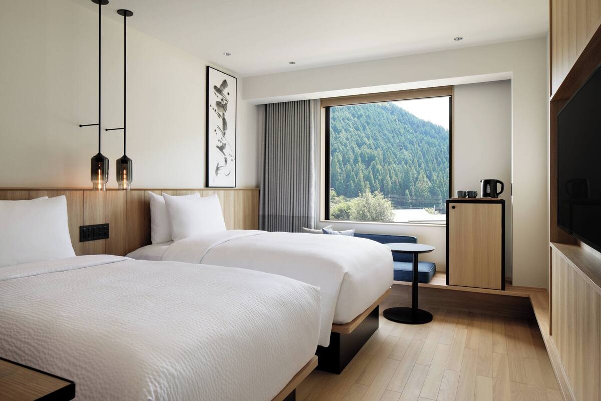 A hotel room with two beds, a large window with a mountain view, a small table with a chair, and a TV on the wall.