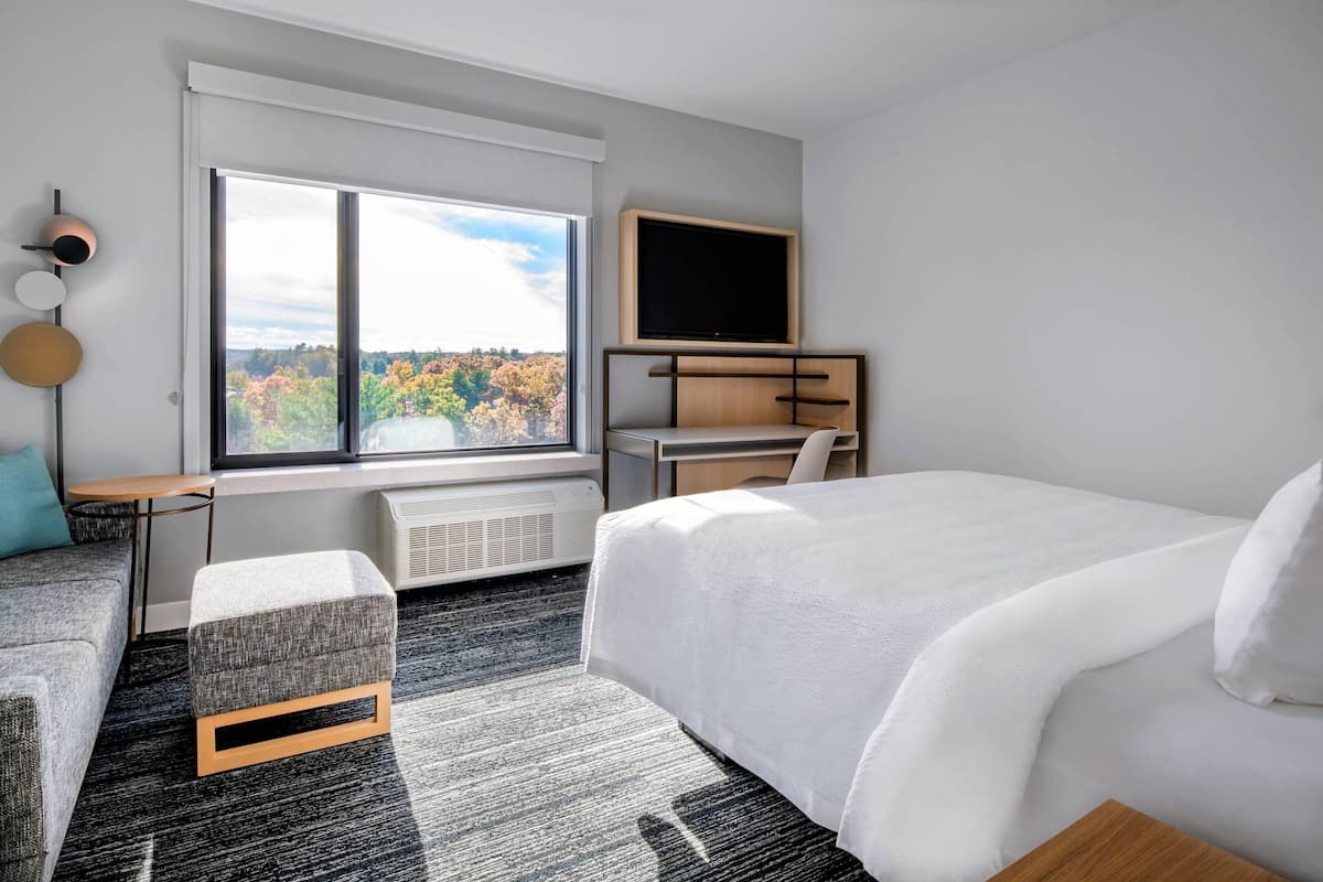 A modern hotel room with a large window, a bed, a desk, and a TV.