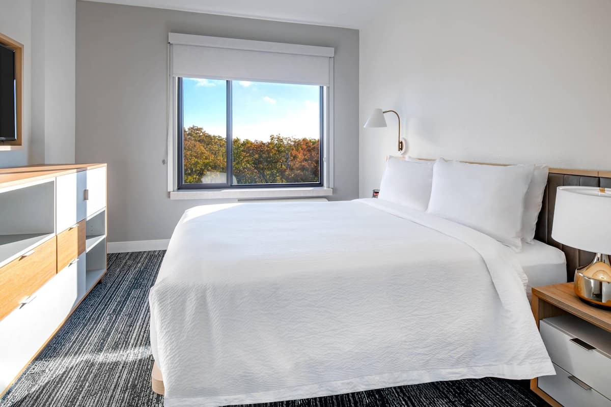 A hotel room with a large bed, a nightstand with a lamp, a dresser, and a window with a view of trees.