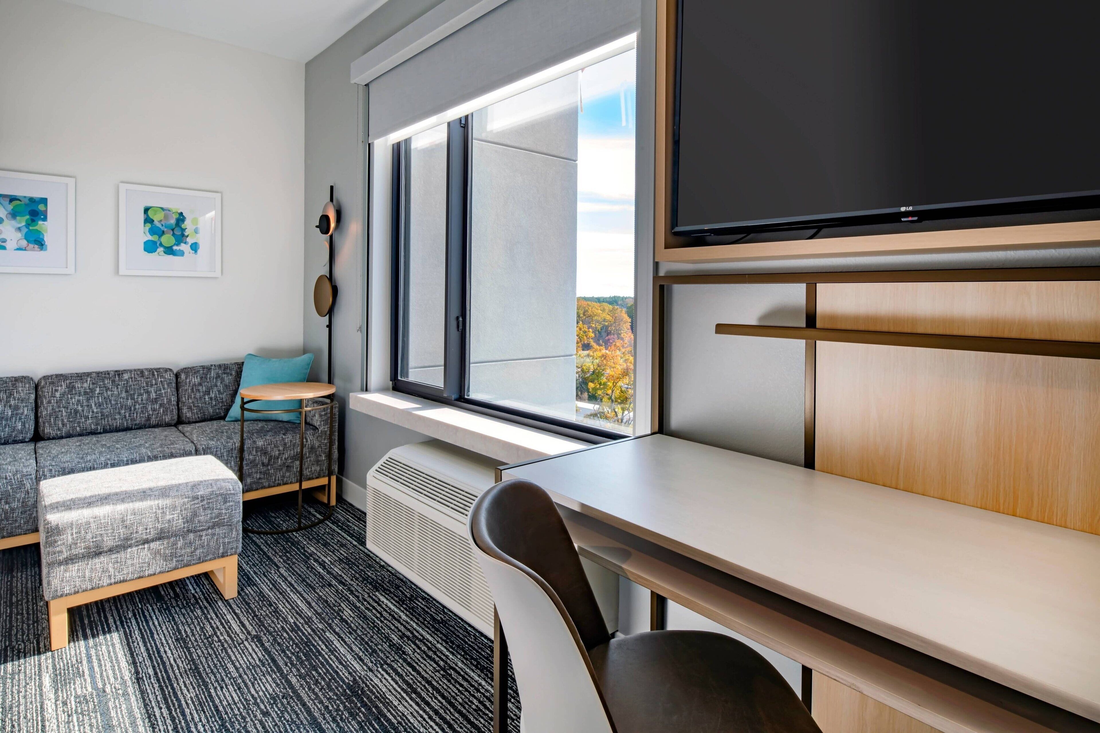 Una habitación de hotel moderna con un sofá gris, una pequeña mesa redonda, un escritorio con una silla, un televisor de pantalla plana y un amplio ventanal con vistas a árboles.
