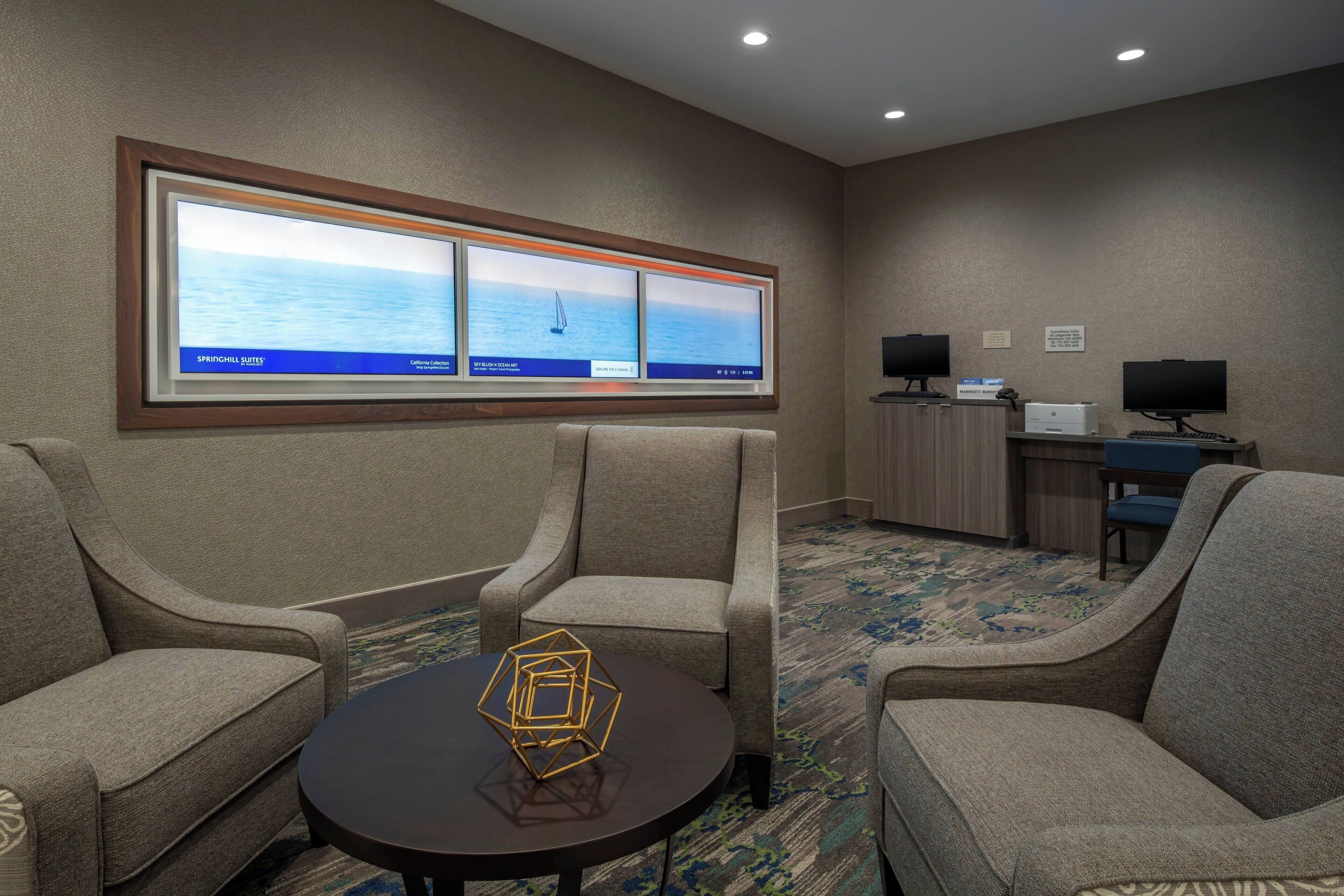 Habitación de hotel con dos sillones, una mesita con un objeto geométrico, un ventanal con vistas al mar y un escritorio con una computadora y un monitor.