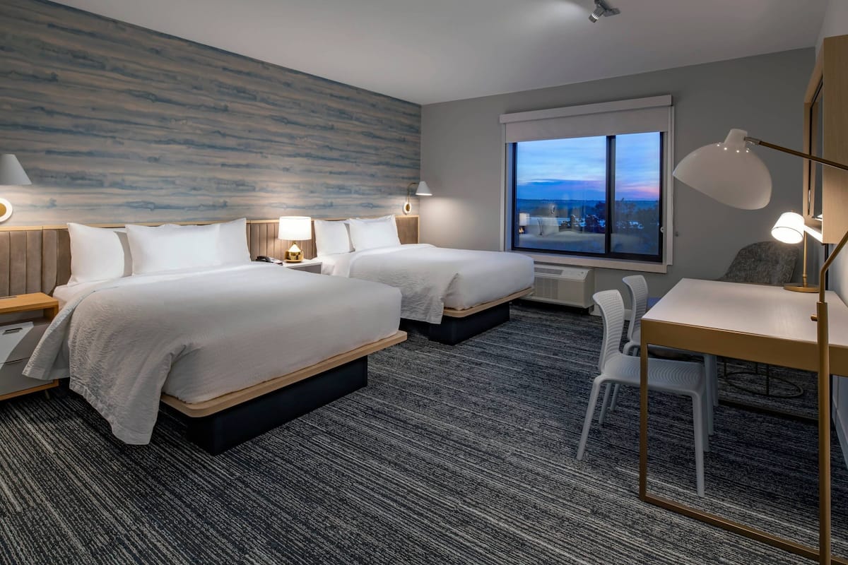 A hotel room with two beds, a desk, and a chair. There is a large window with a view of the cityscape.