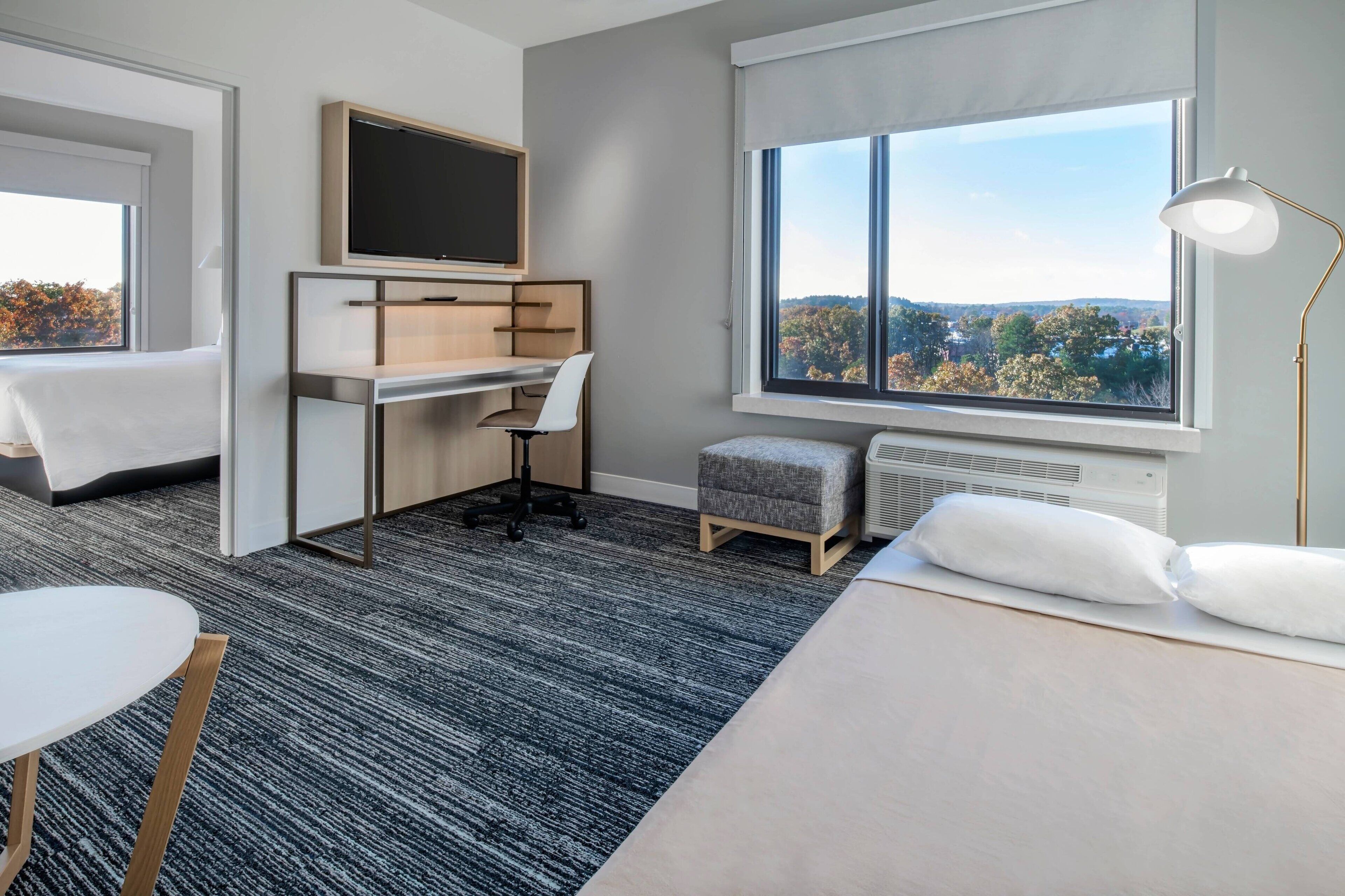 A modern hotel room with a bed, a desk, a chair, a TV, a window with a view, and a lamp.