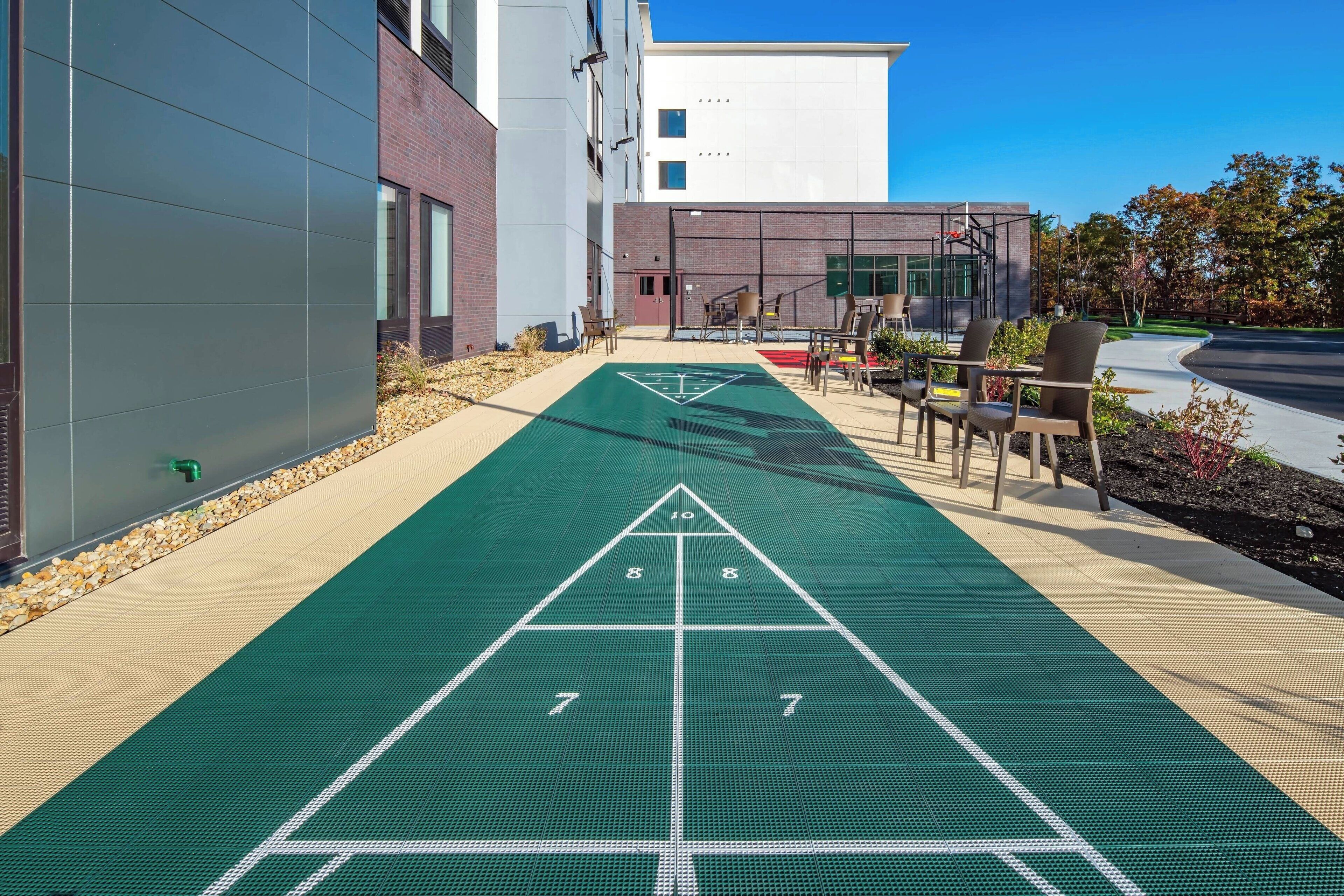 A shuffleboard court adjacent to a building with a modern design, outdoor seating, and a clear sky.