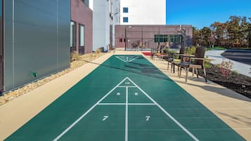 A shuffleboard court adjacent to a building with a modern design, outdoor seating, and a clear sky.