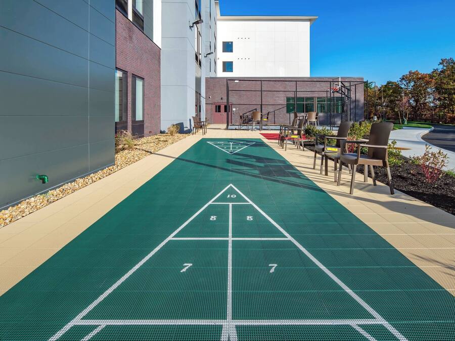 A shuffleboard court adjacent to a building with a modern design, outdoor seating, and a clear sky.
