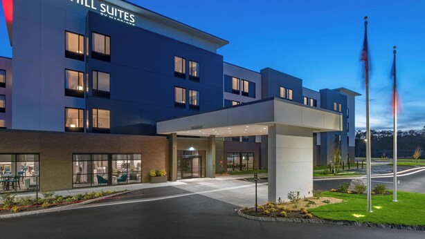 A modern hotel with a flat roof, large windows, and a well-lit entrance. - TownePlace Suites by Marriott Wrentham Plainville (Plainville)