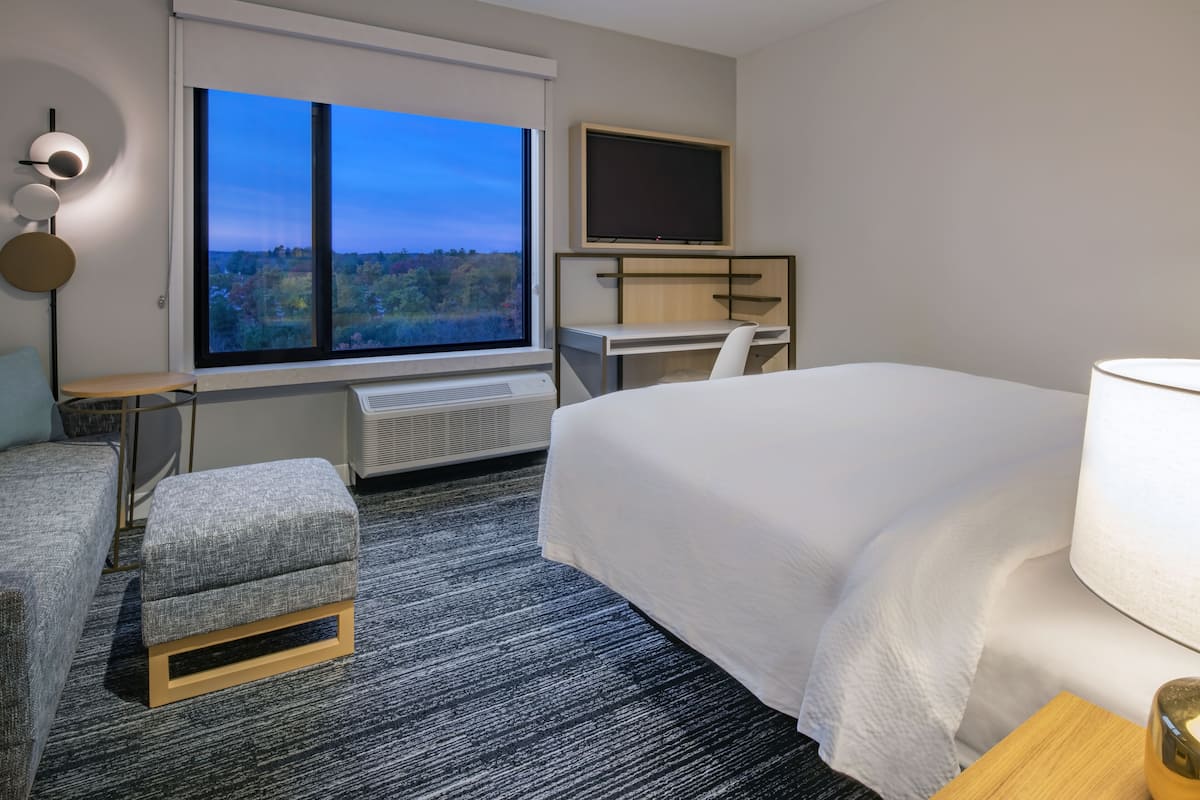 A modern hotel room with a large bed, a desk, a chair, a small table, a TV, and a large window offering a scenic view.