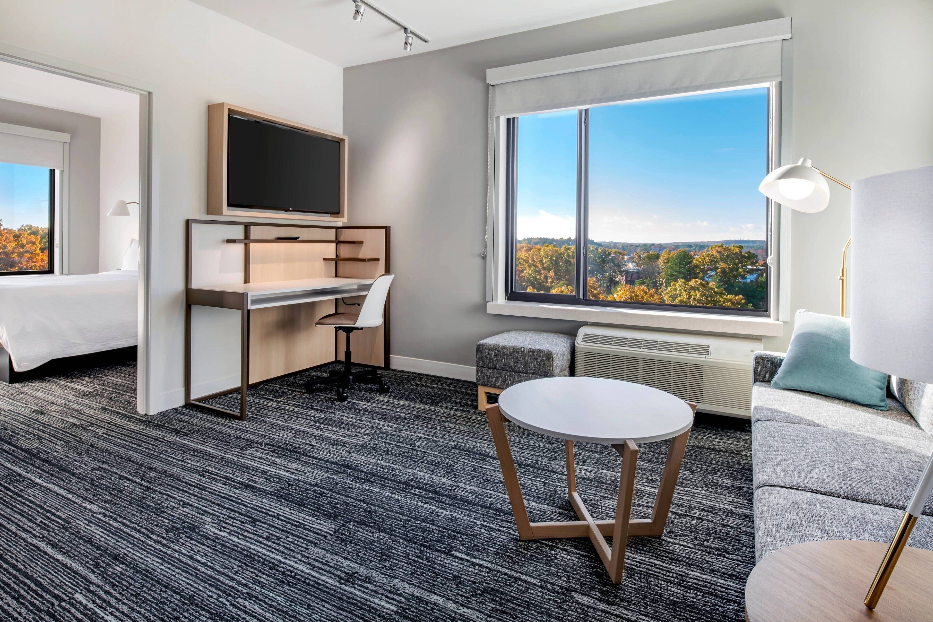 A modern hotel room with a bed, a desk with a TV, a large window with a city view, and a cozy seating area.