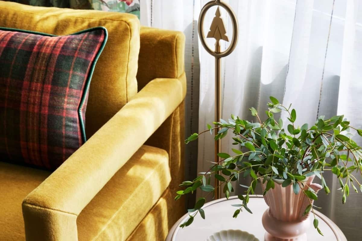 Un canapé de couleur moutarde agrémenté d’un coussin à carreaux, une petite table blanche sur laquelle sont posés un vase rose et une plante verte, et un panneau décoratif en métal.