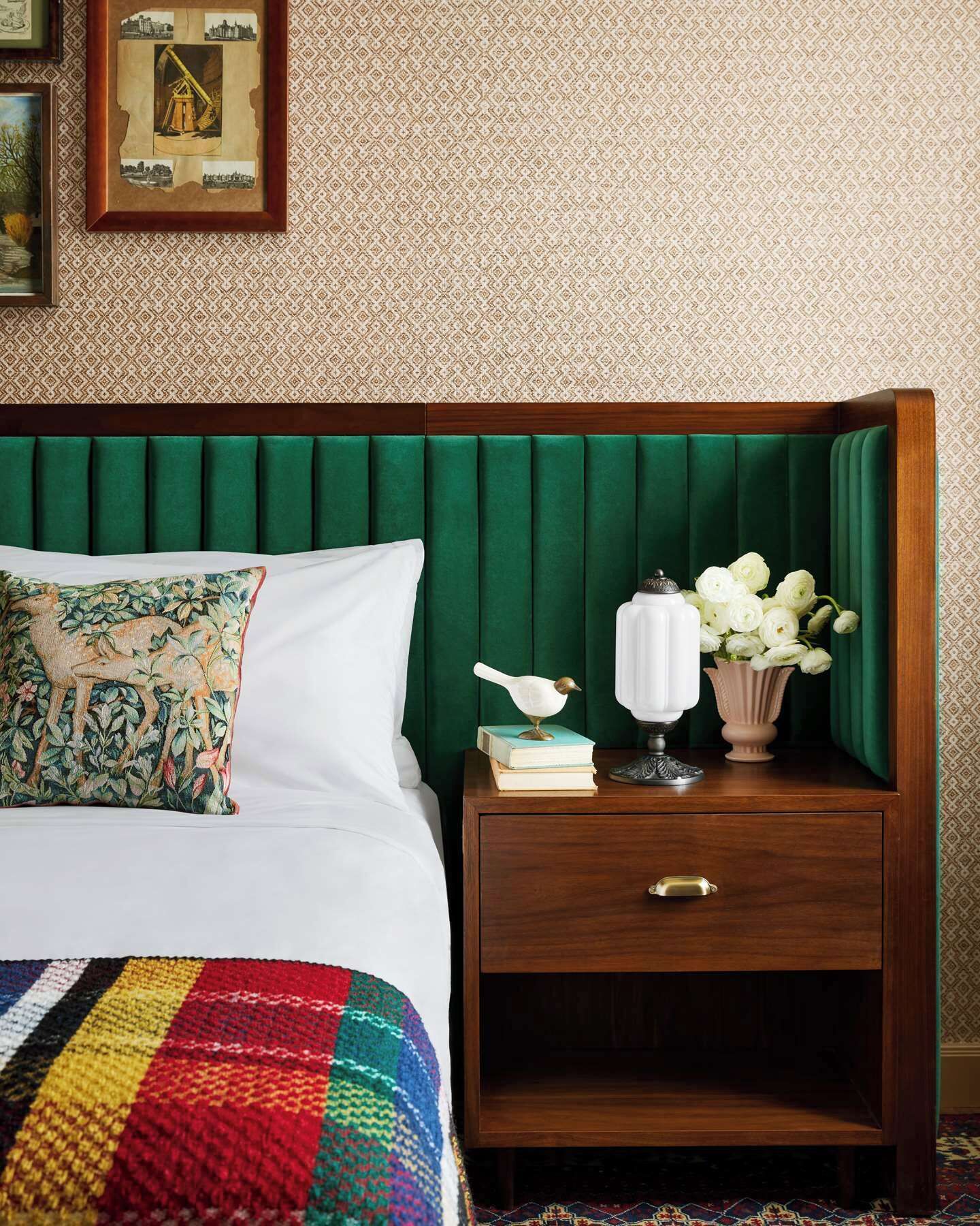 Una cama bien hecha con un edredón colorido, un cabecero tapizado en verde, una mesita de noche con una lámpara blanca, una figurita de pájaro y un jarrón con flores blancas.