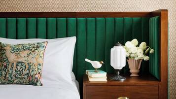 Una cama bien hecha con un edredón colorido, un cabecero tapizado en verde, una mesita de noche con una lámpara blanca, una figurita de pájaro y un jarrón con flores blancas.