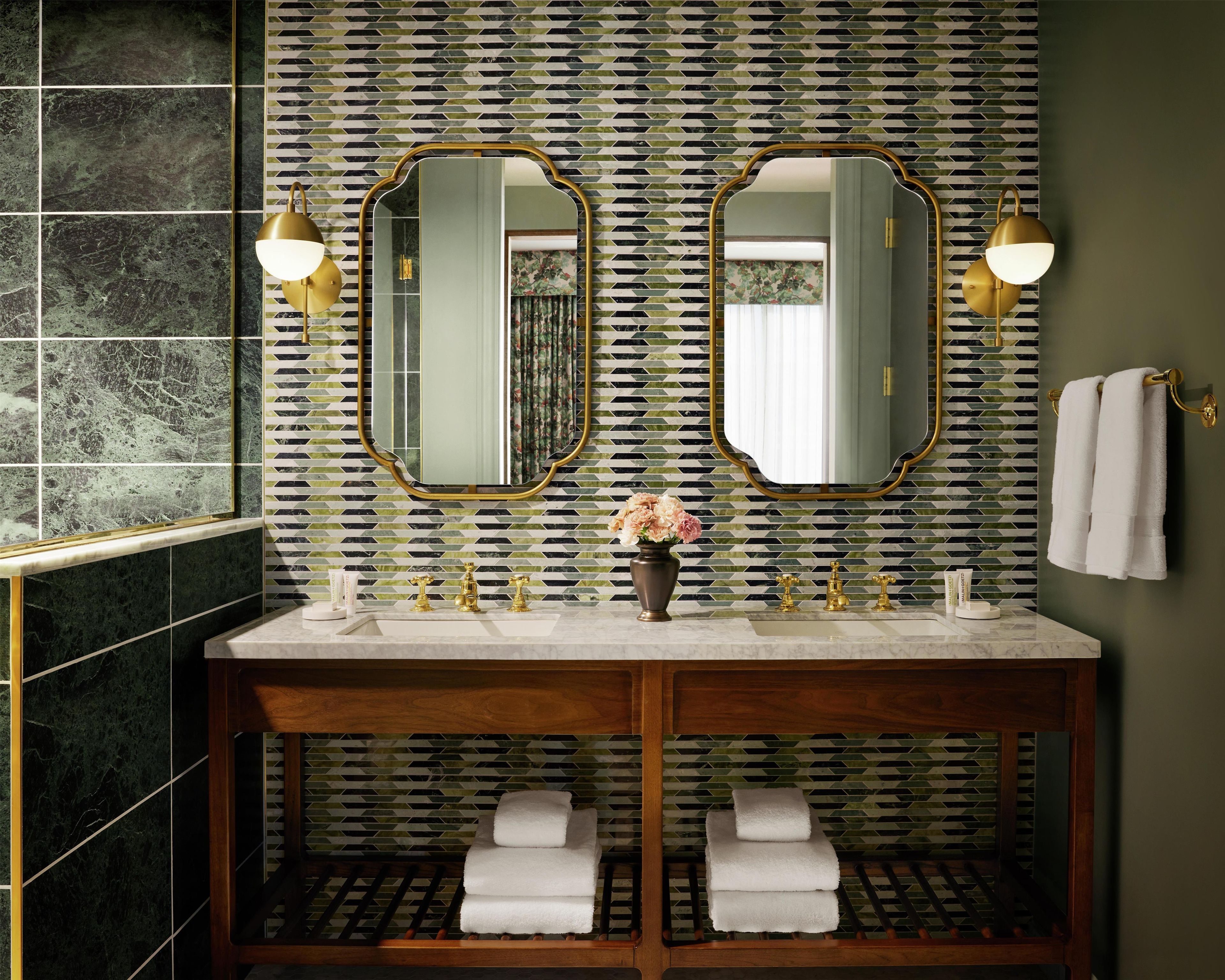 A bathroom with a dual vanity, two mirrors with gold frames, a vase with flowers, and a towel rack with towels.