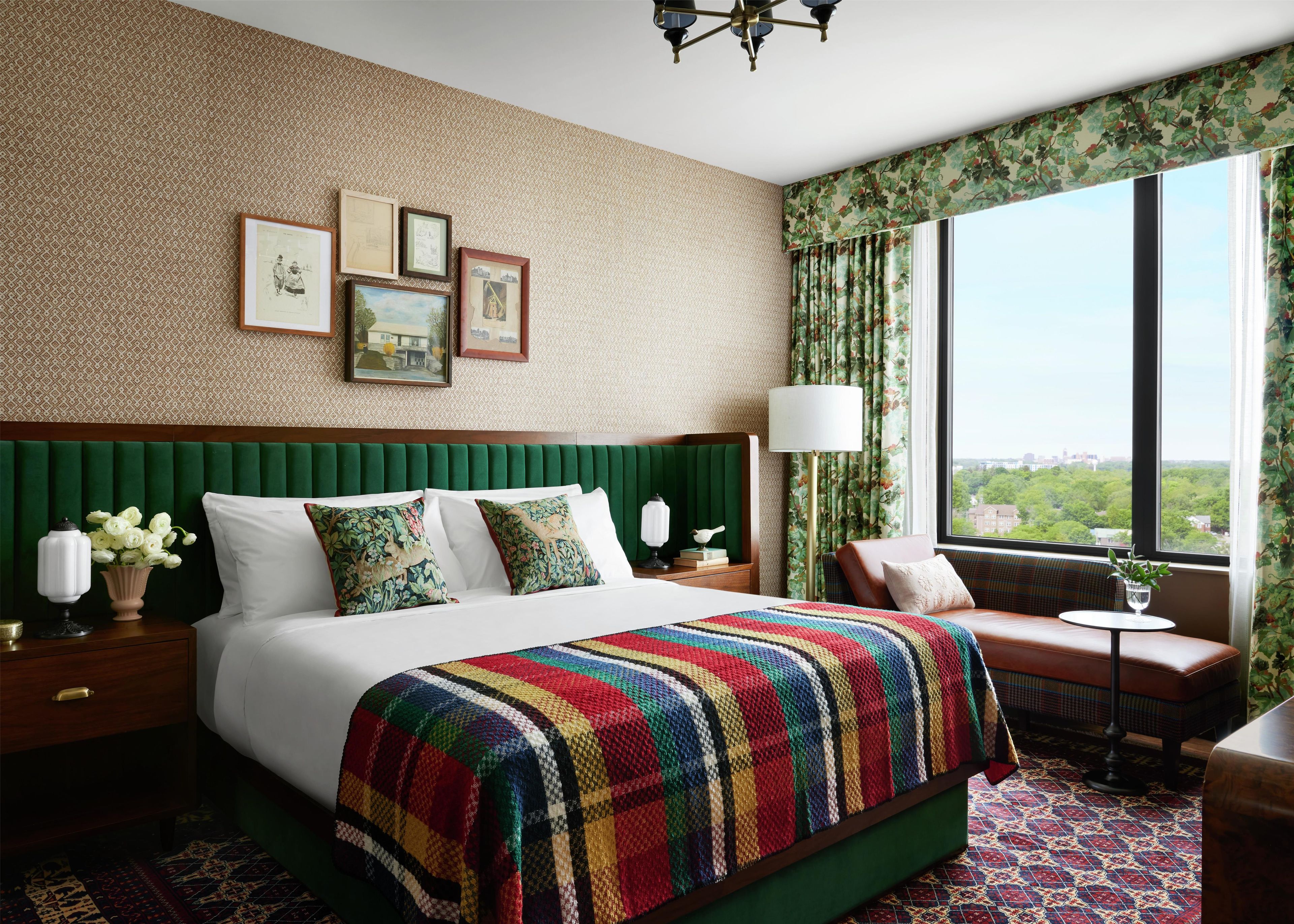 Habitación de hotel con una cama grande, cabecera tapizada de verde, vistas de la ciudad y una ventana con cortinas florales.
