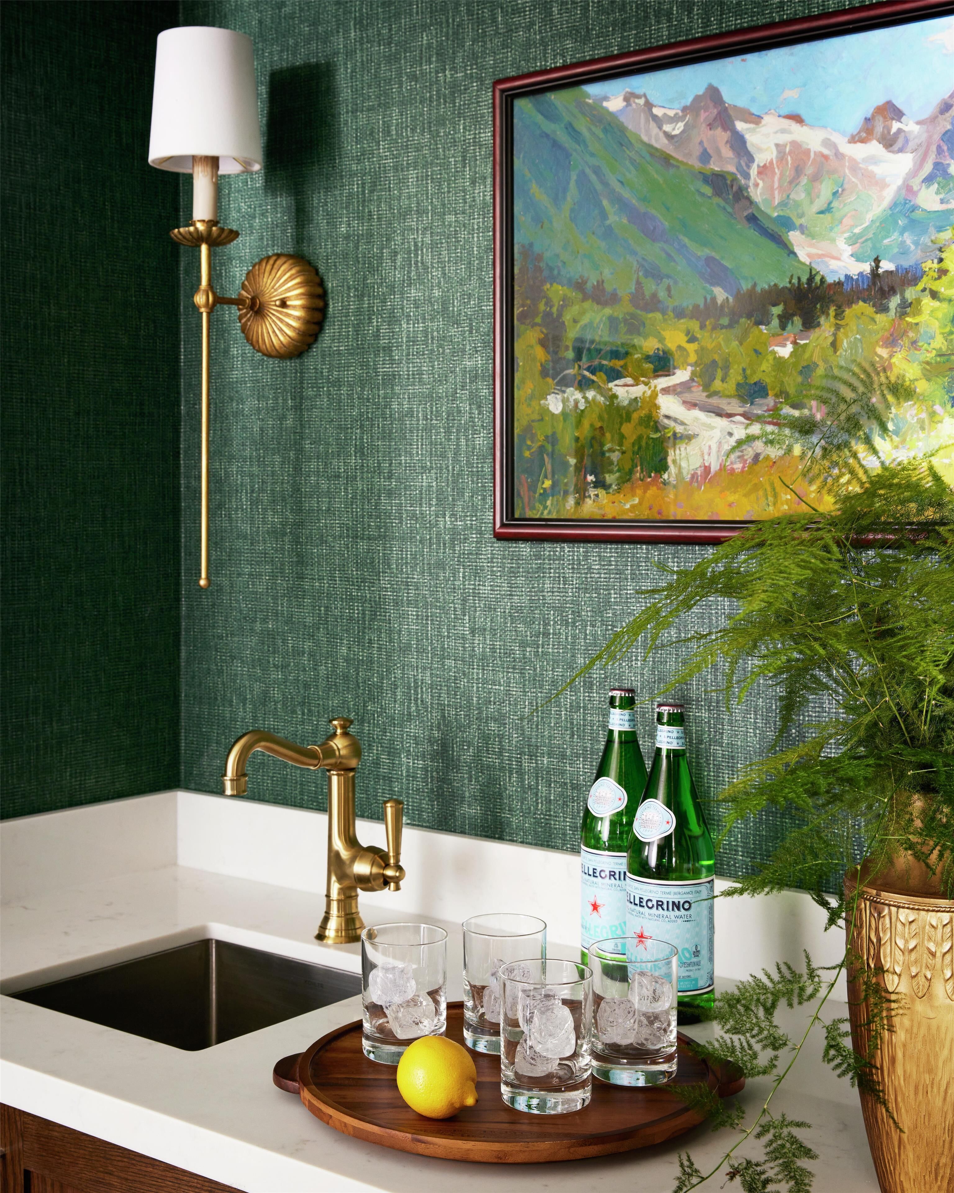 A kitchen counter with a sink, a gold faucet, and a tray with glasses and a lemon. There are two bottles of water and a potted plant.