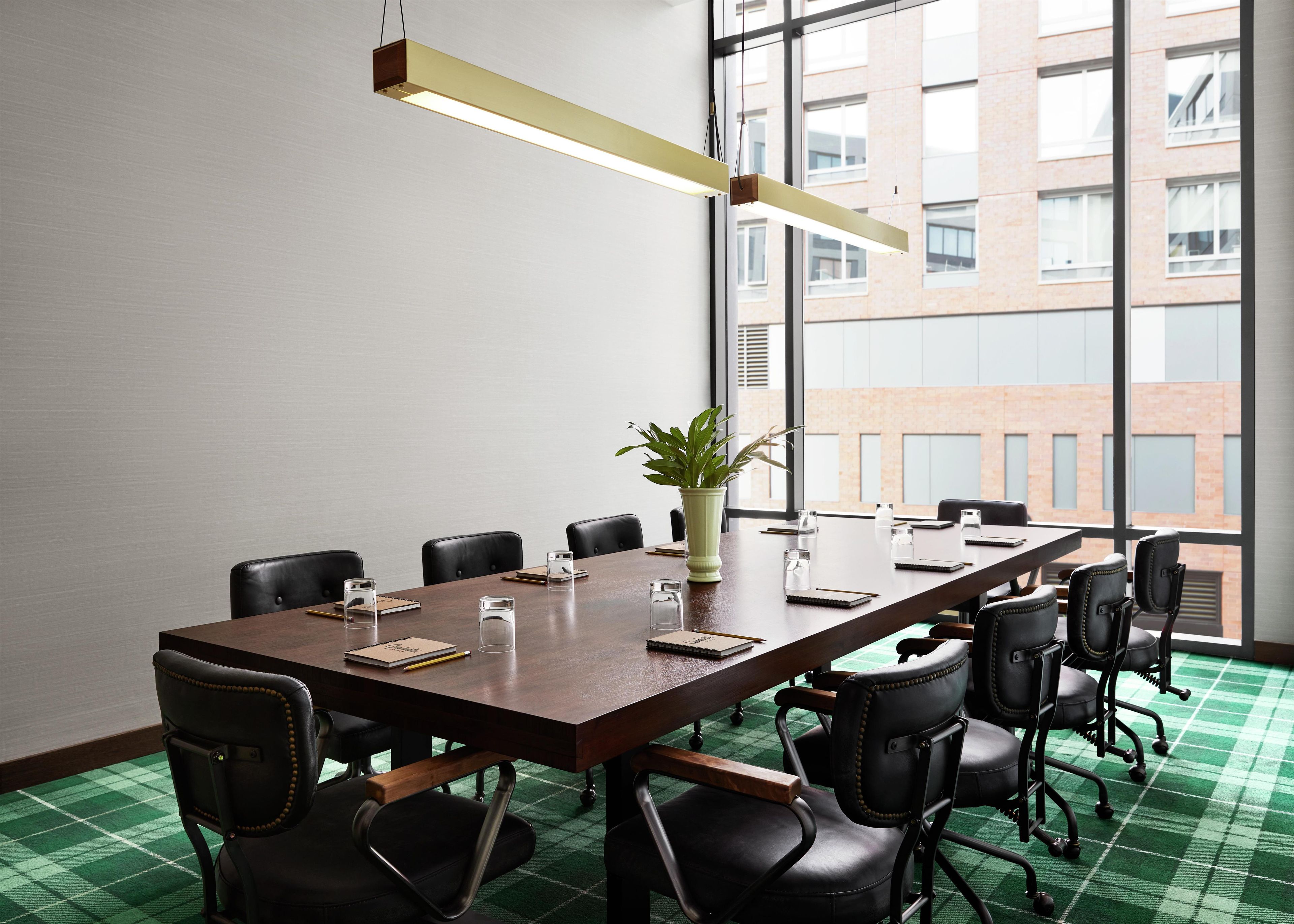 Un salón de conferencias con una larga mesa, sillas negras, una planta en maceta y una lámpara colgante moderna.