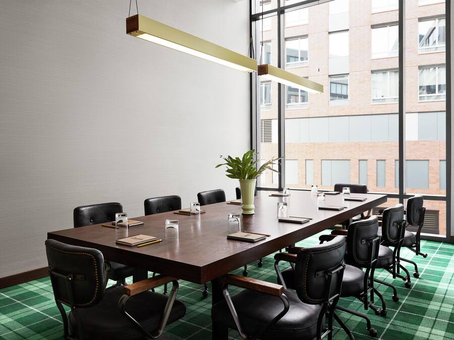 Un salón de conferencias con una larga mesa, sillas negras, una planta en maceta y una lámpara colgante moderna.