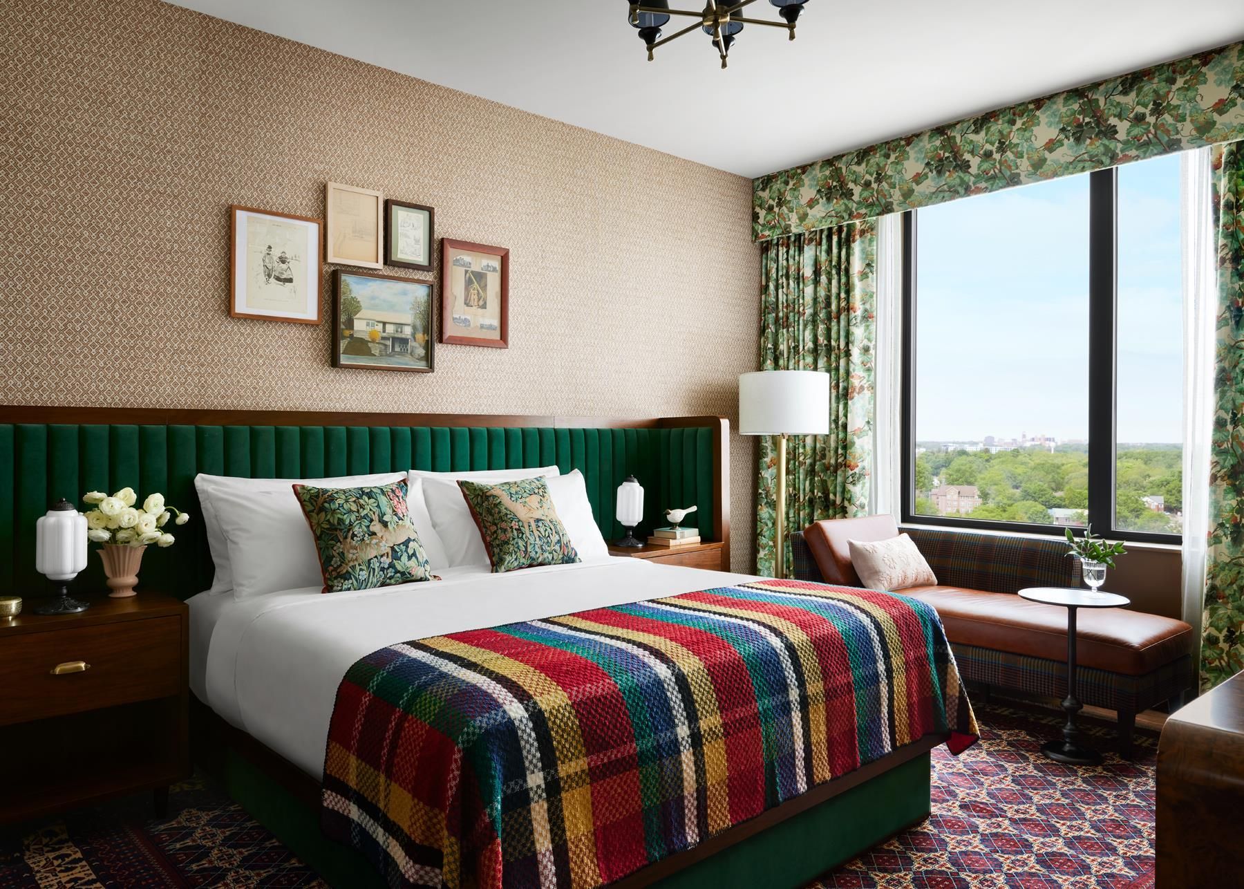 A hotel room with a large bed, a green upholstered headboard, a window with a view, and a patterned carpet.