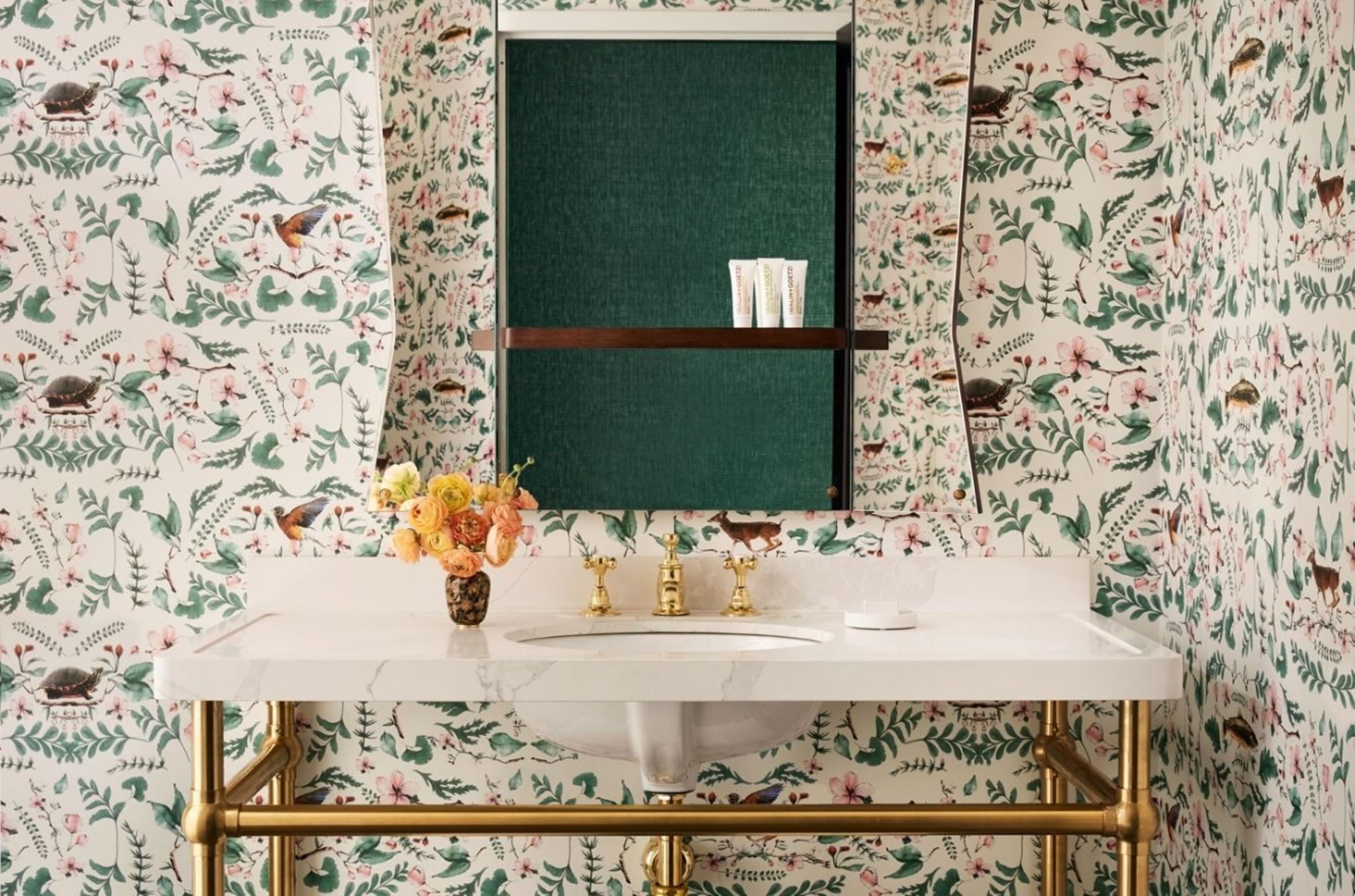 Ein Badezimmer mit floraler Tapete, einem weißen Waschbecken, goldenen Armaturen und einem grünen Spiegel.