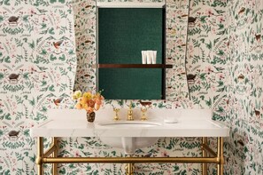 A bathroom with a floral wallpaper, a white sink, gold fixtures, and a green mirror.