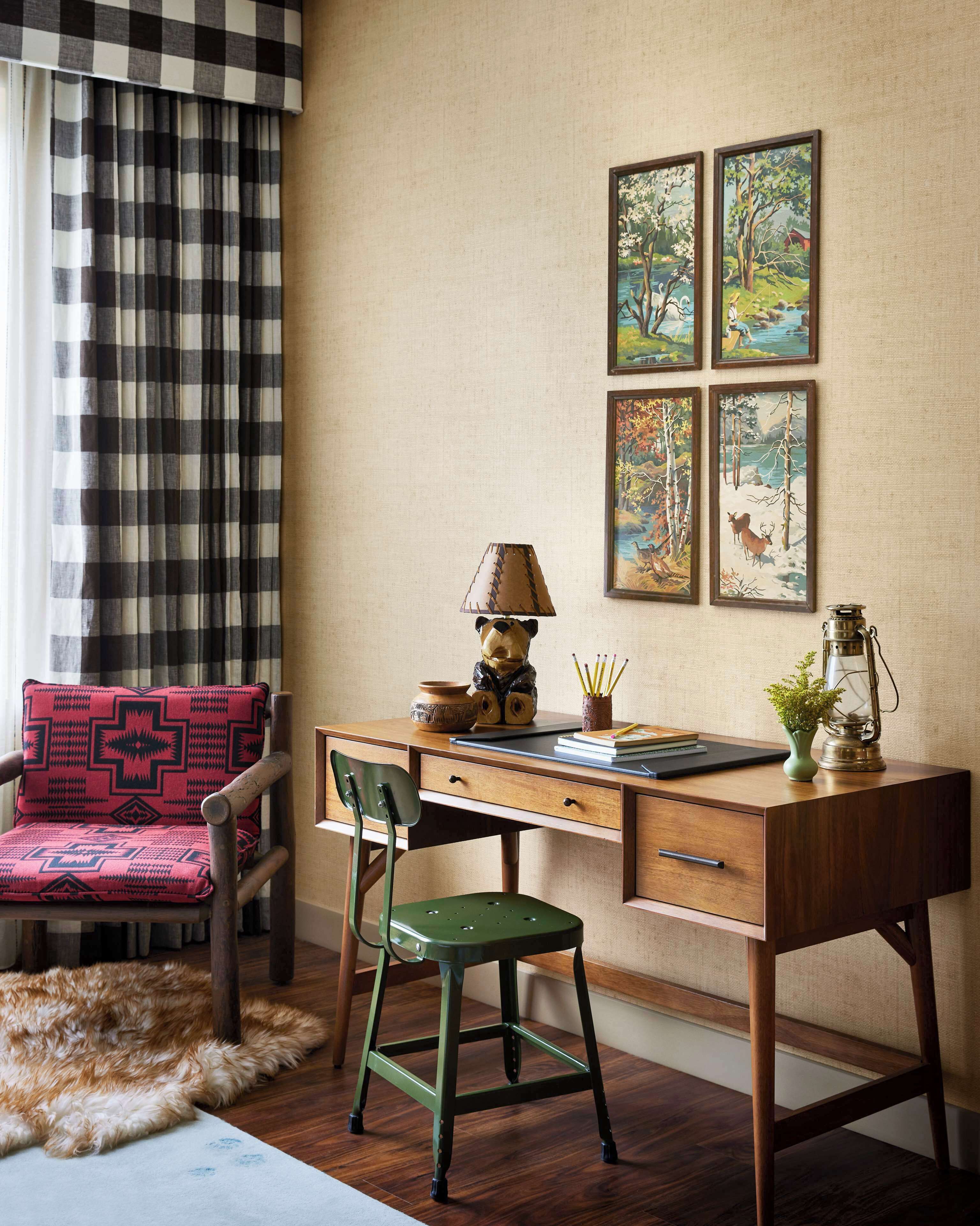 A room with a wooden desk, a chair, a lamp, and four framed paintings on the wall.